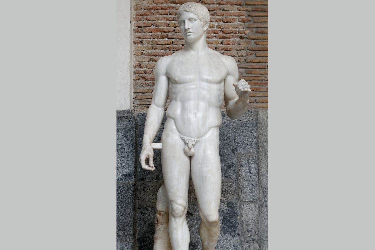 Essa transformação não se limitava à escultura. A própria filosofia começava a imaginar que o universo obedecia a relações proporcionais. Como observa Umberto Eco em História da Beleza, Pitágoras sustentava que o princípio de todas as coisas era o número. Diante do infinito, os pitagóricos buscavam na proporção uma forma de tornar o mundo compreensível. Talvez por isso a simetria tenha se tornado um dos grandes ideais da arte grega clássica. Em Doríforo, do século V a.C., essa confiança atinge um de seus pontos mais altos. O peso do corpo se desloca suavemente sobre uma das pernas, enquanto o tronco acompanha discretamente o movimento. A harmonia surge menos da rigidez do que de uma contenção perfeita. O Doríforo que hoje contemplamos, entretanto, já pertence também à história das perdas da Antiguidade. Muitas esculturas gregas chegaram até nós por meio de cópias romanas, já que os originais em bronze desapareceram ao longo do tempo