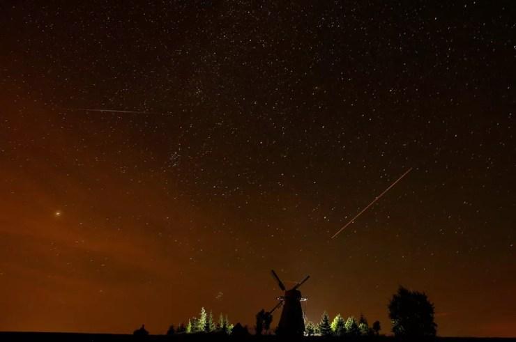 Traços de um meteoro, à esquerda, e de um avião, à direita e mais avermelhado, riscam o céu em fotografia de longa exposição no céu de Ptich, Belarus, durante a chuva de meteoros Perseidas (Foto: Vasily Fedosenko/Reuters)
