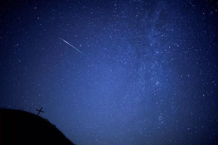 Meteoro corta o céu, com a Via Láctea vista à direita no céu e uma cruz à esquerda sobre a colina Leeberg, em Grossmugl, na Áustria, durante as Perseidas (Foto: Heinz-Peter Bader/Reuters)