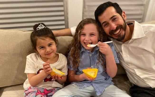 Fotografia do pai, junto com as duas filhas, enquanto as duas crianças comem.
