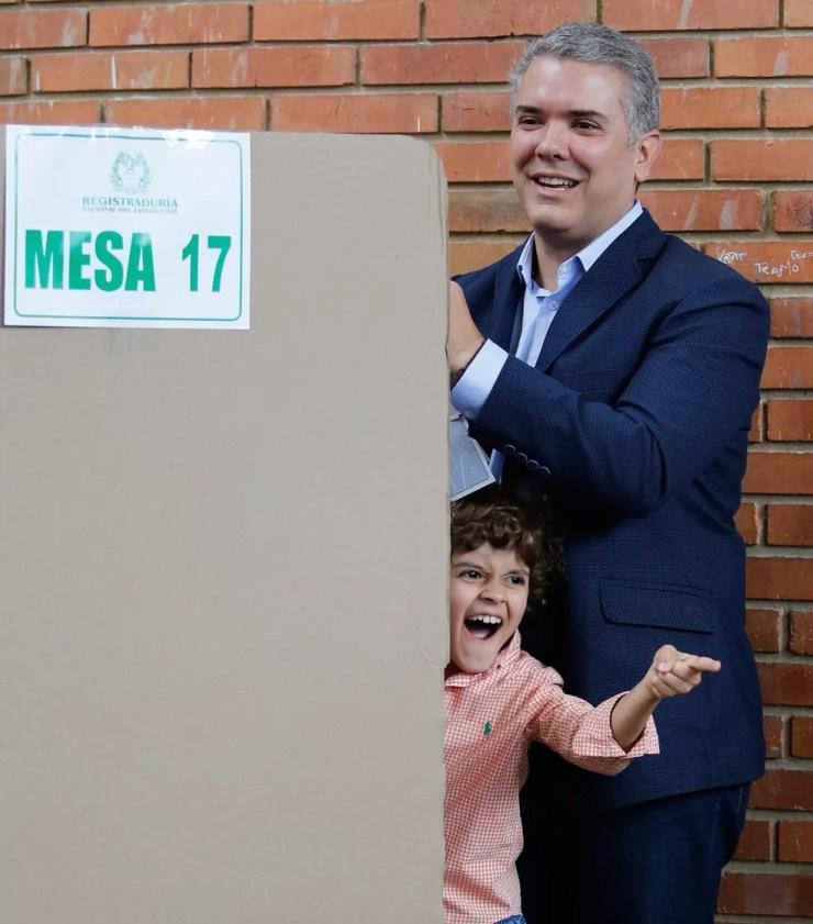 O candidato à Presidência da Colômbia Ivan Duque, do partido Centro Democrático, vota em uma urna em Bogotá enquanto seu filho rouba a cena fazendo caretas para a família e os fotógrafos na sala (Foto: Fernando Vergara/AP)